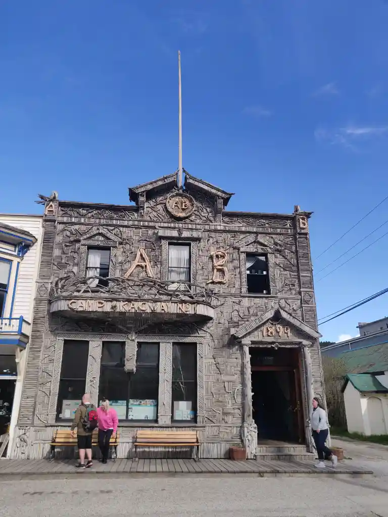 A historic wooden building labeled 'CAMP KAGWAY NO 1' and dated '1899' features intricate twig and branch carvings, with…
