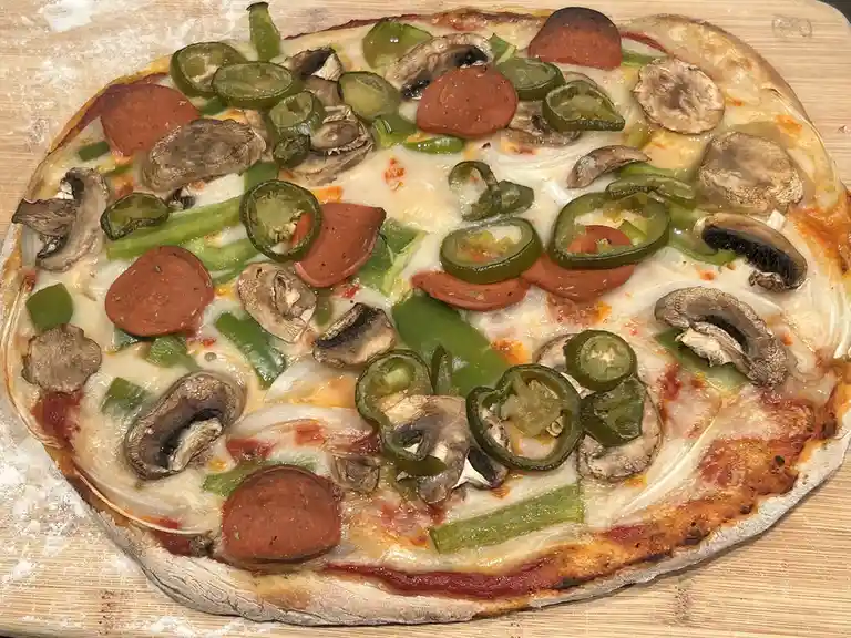 A homemade pizza with pepperoni, sliced jalapeños, mushrooms, and green bell peppers sits on a wooden cutting board dusted…