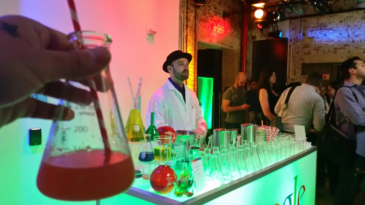 A person holds a beaker with red liquid and a striped straw in the foreground, while a bartender in a white lab coat and…