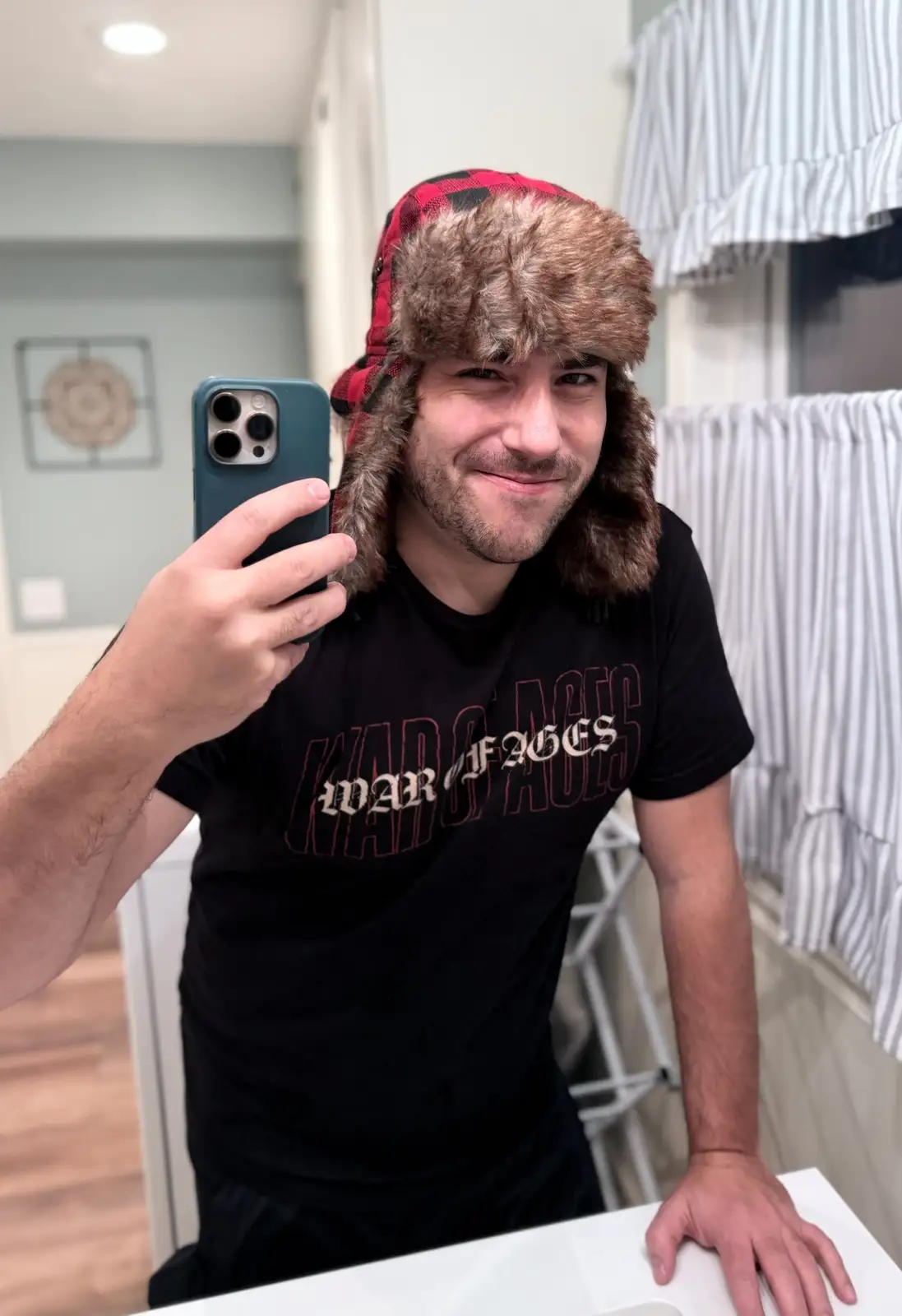 A man takes a mirror selfie holding a teal phone, wearing a red plaid fur-trimmed hat and a black t-shirt with 'WAR OF AGES'…