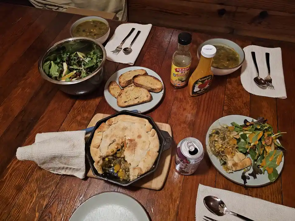 A wooden dining table is set with a cast-iron skillet containing a partially eaten savory pie, surrounded by plates of…