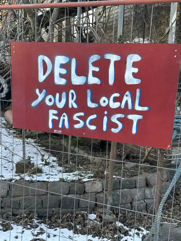A red wooden sign with white spray-painted text reading 'DELETE YOUR LOCAL FASCIST' is mounted on a wire fence in a snowy…