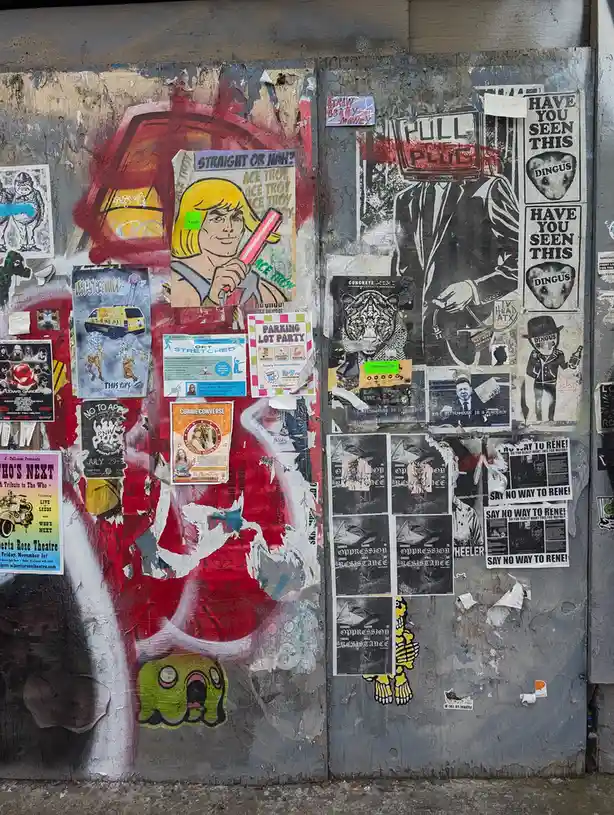 A weathered wall covered in overlapping posters, flyers, and graffiti, including a prominent 'HAVE YOU SEEN THIS DINGUS'…
