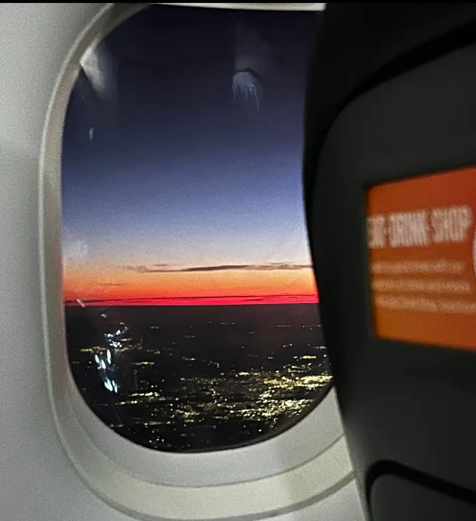 A view from an airplane window shows a vibrant sunset or sunrise over a cityscape with lights reflecting on water, framed by…