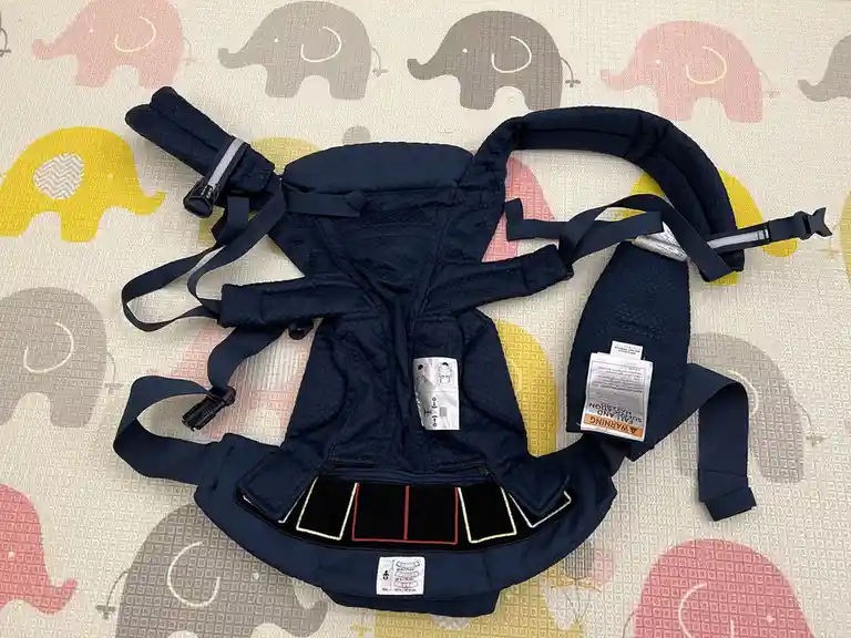 A navy blue baby carrier lies unfolded on a colorful mat with cartoon elephants, showing its straps, buckles, and warning…