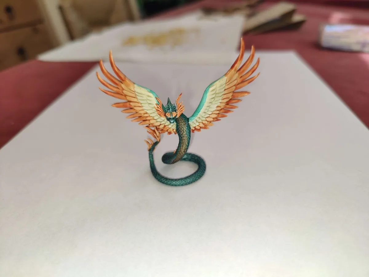 A painted miniature dragon figurine with teal scales, orange-tipped wings, and a coiled tail stands on a white surface. The…