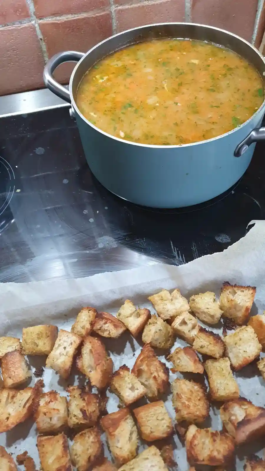A pot of orange-colored soup with green herbs sits on a black stovetop, next to a baking sheet lined with parchment paper…