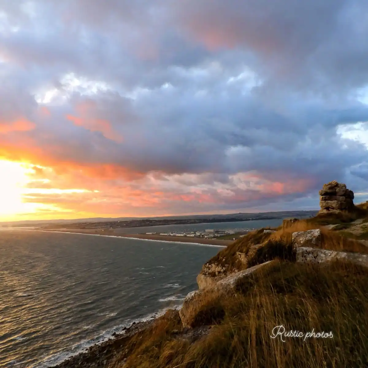 A scenic coastal sunset photo shows a dramatic sky with orange and pink clouds over a choppy sea, a distant shoreline, and a…