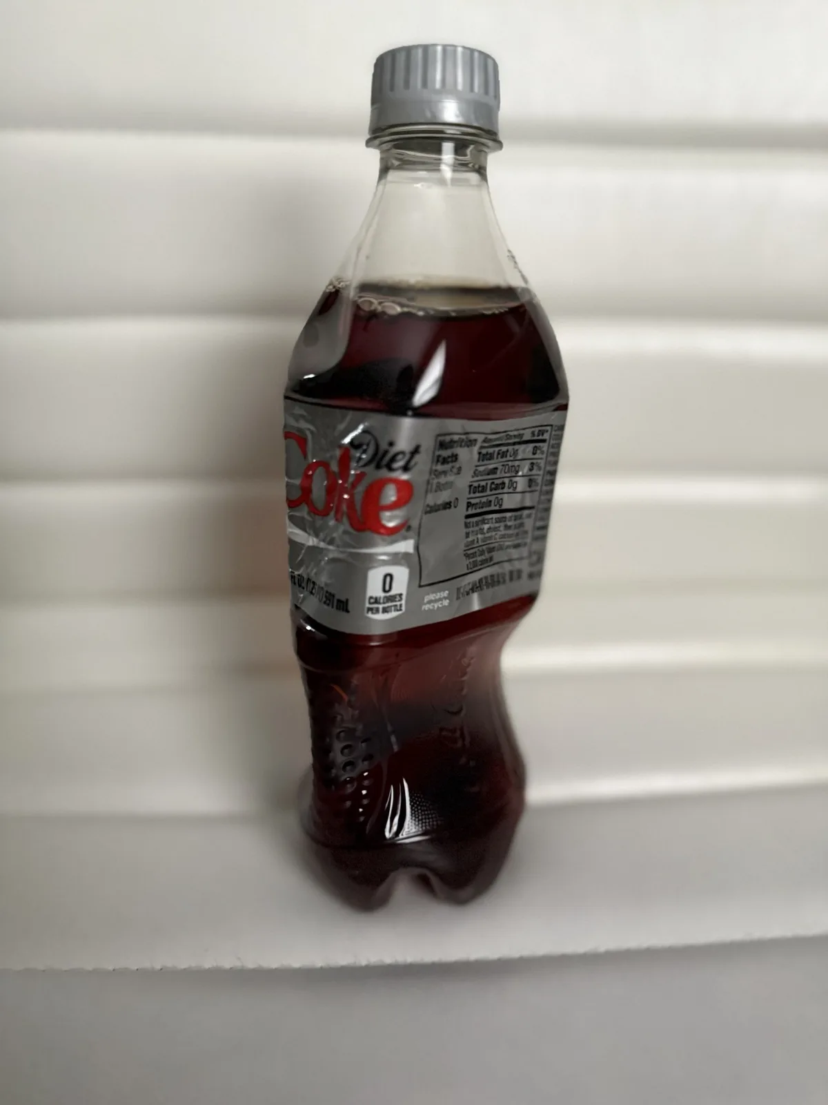 A plastic bottle of Diet Coke with a silver cap sits on a white surface, showing its nutrition label and '0 CALORIES PER…