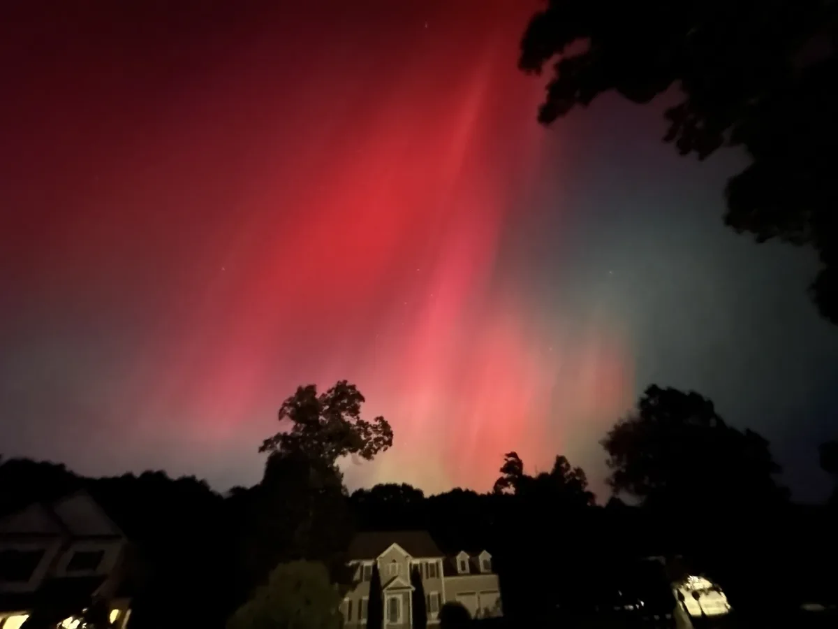 A nighttime photo shows a vivid red aurora borealis streaking across the sky above silhouetted suburban houses and trees…