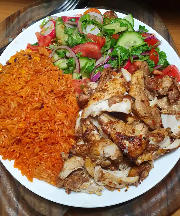 A white plate holds a large serving of vibrant orange rice, shredded grilled chicken, and a fresh salad with cucumbers…