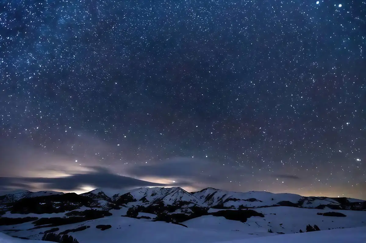 A breathtaking night sky filled with countless stars and subtle nebulae stretches above a snow-covered mountain range. The…
