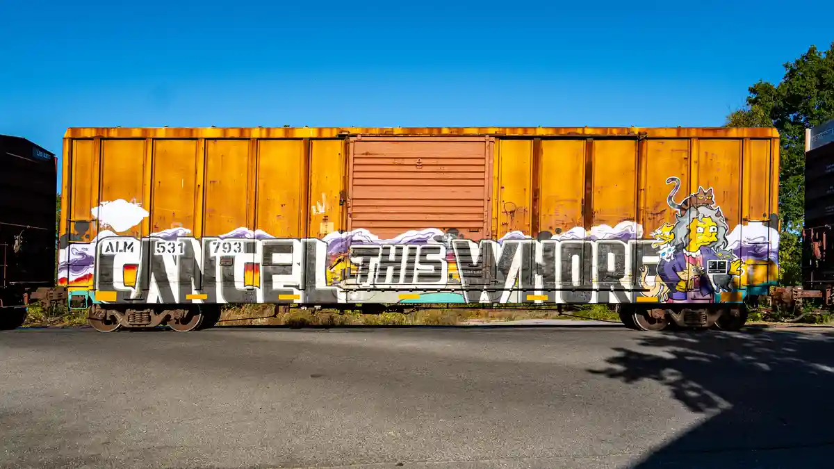 A rusty orange freight train car is covered in graffiti, prominently featuring the phrase 'CANCEL THIS WHORE' in large…