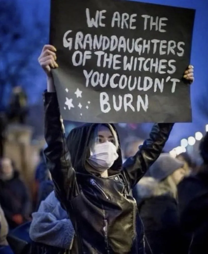 A person wearing a face mask and a black hooded jacket holds up a black protest sign with white handwritten text that reads…