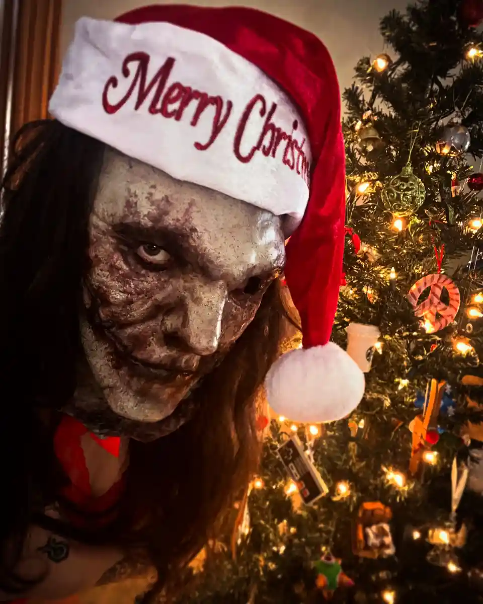 A person with a zombie-like, decayed face and long dark hair wears a red and white 'Merry Christmas' Santa hat while…