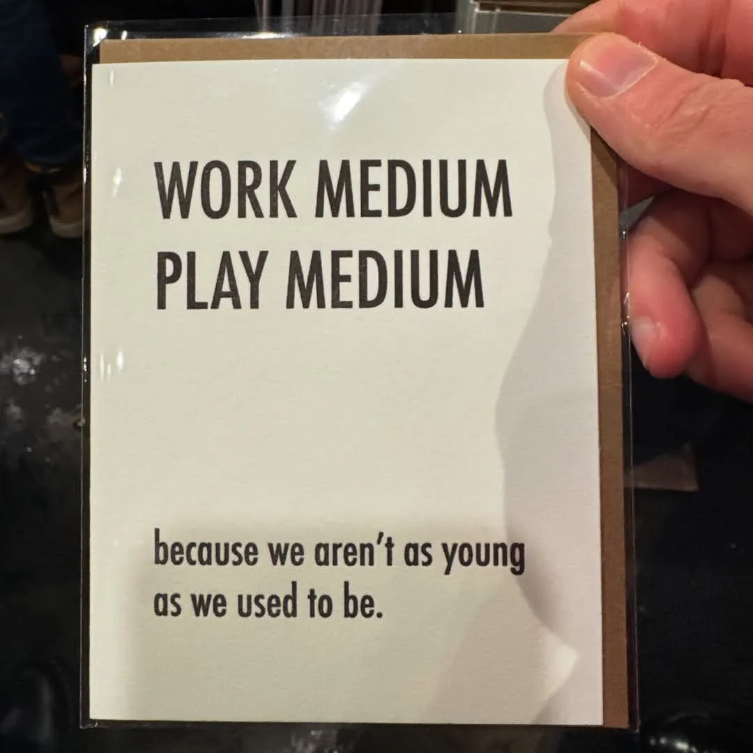 A hand holds a white card with brown borders, displaying the text 'WORK MEDIUM PLAY MEDIUM' in large bold letters, followed…