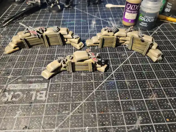 Three miniature wargaming barricades made of sandbags and wooden crates sit on a gridded painting mat, each with tiny…