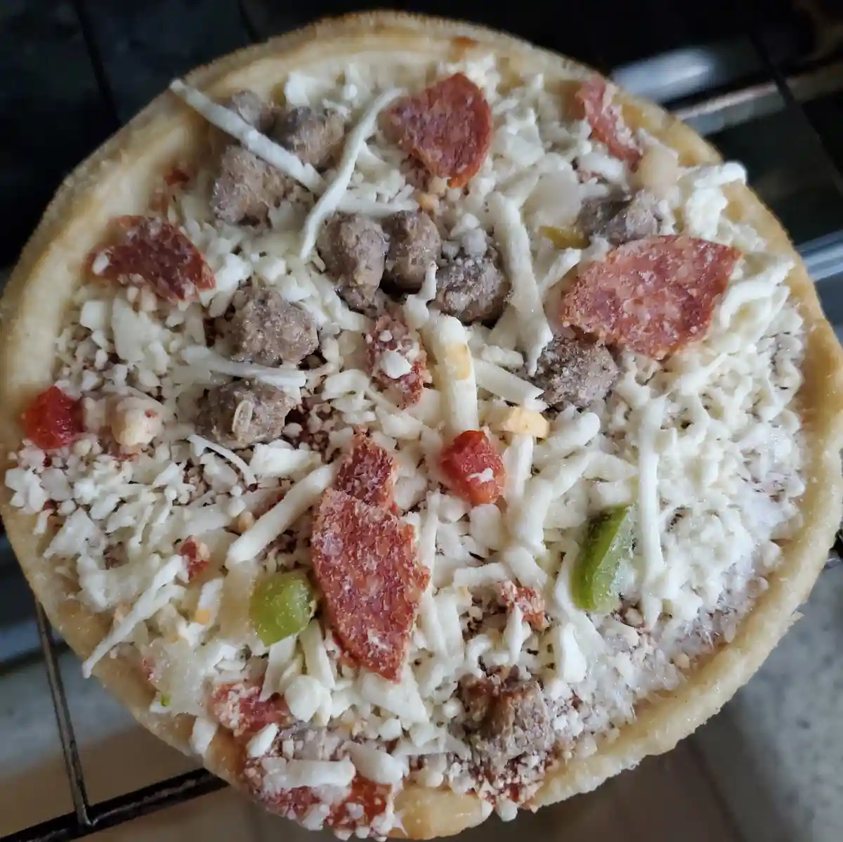A close-up photo of an uncooked pizza with a thick crust, covered in shredded white cheese, crumbled sausage, pepperoni…