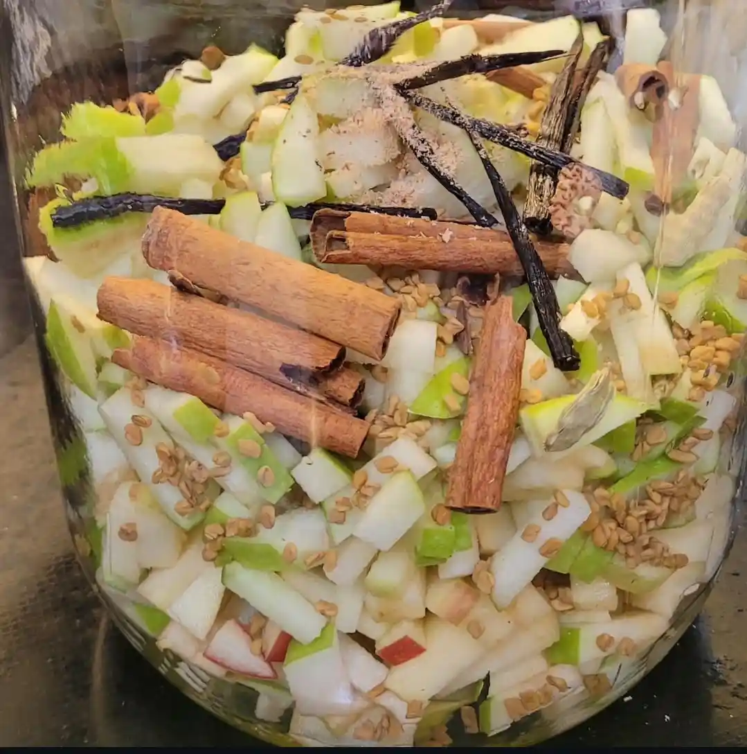 A clear glass jar filled with chopped green and white apples, whole cinnamon sticks, vanilla beans, and small seeds, with a…
