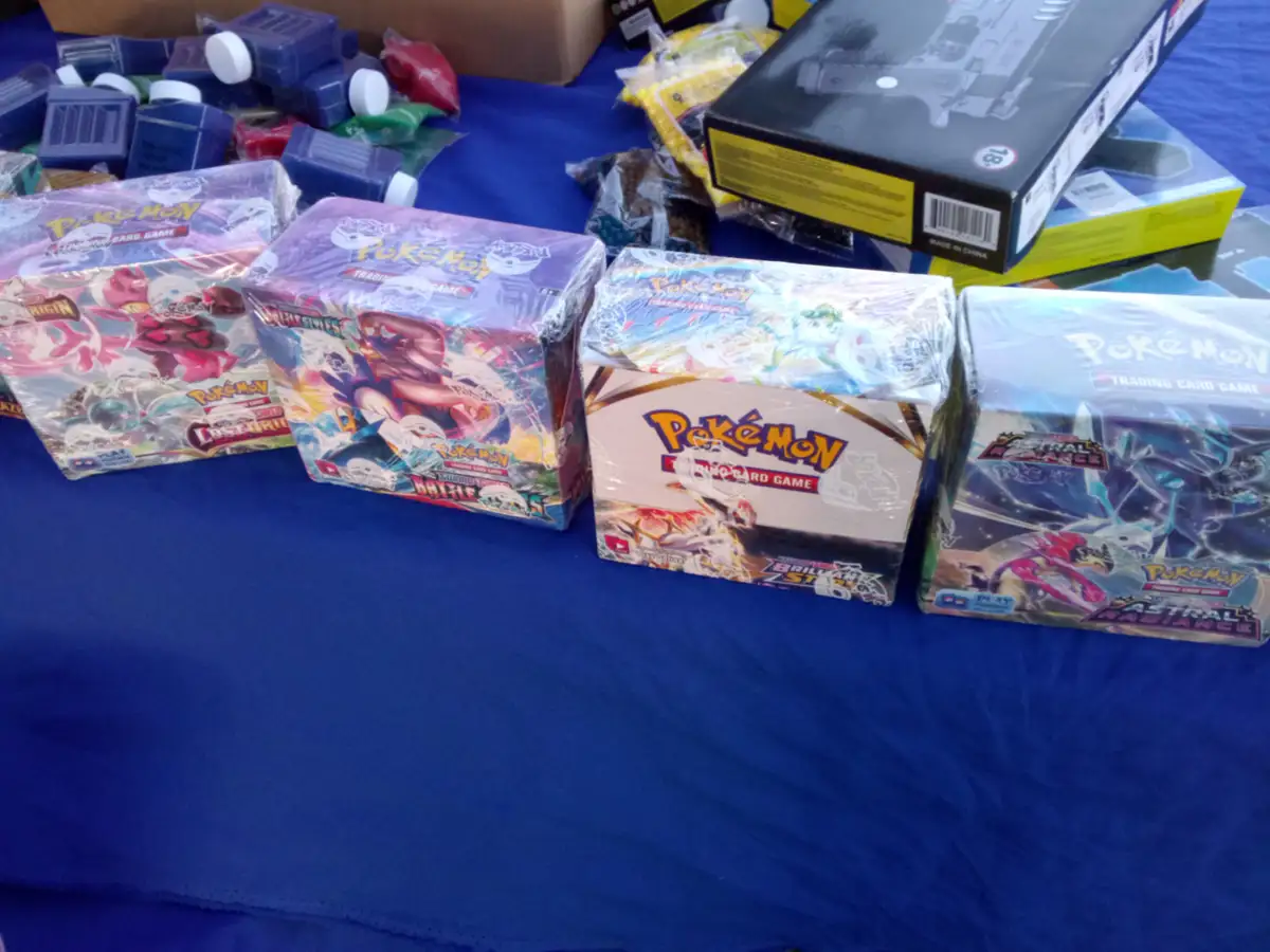 A blue table is covered with several unopened Pokémon Trading Card Game booster boxes, including sets like 'Astral Radiance'…