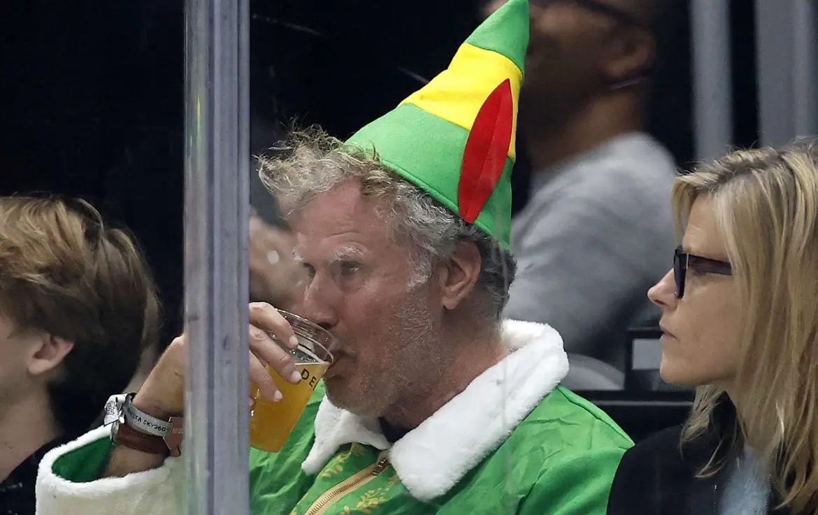 A man with gray hair wearing a green, yellow, and red striped elf hat and a matching green jacket with white fur trim is…