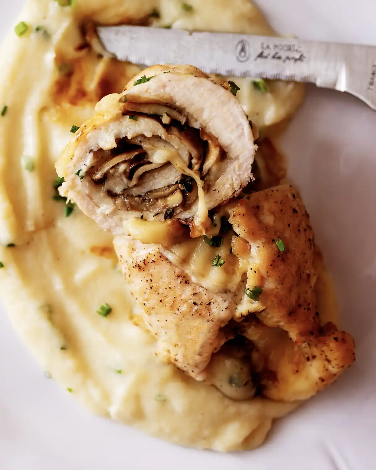 A close-up photo of a sliced chicken roll filled with mushrooms and cheese, served on creamy mashed potatoes with a knife…