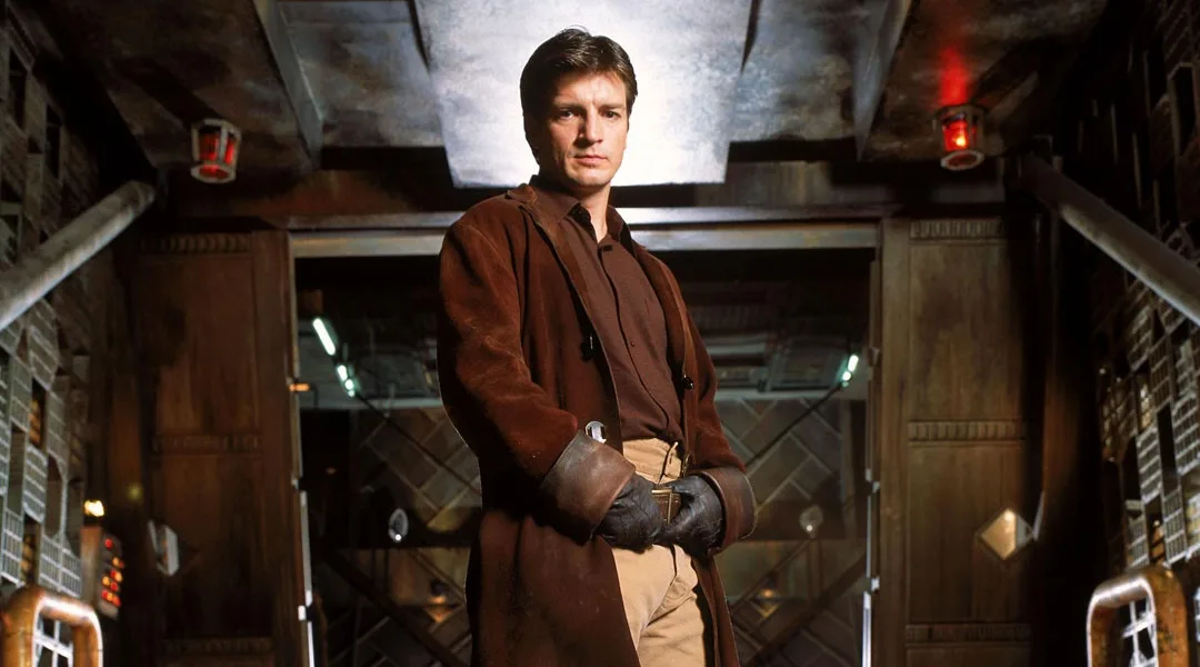 A man in a brown coat and gloves stands in a dimly lit, industrial-style corridor with red emergency lights. The overlaid…