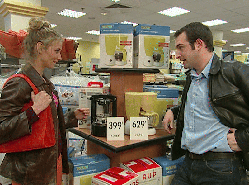 A man and woman are in a store aisle with coffee makers priced at 399 and 629. The woman hands the man a box, then walks…