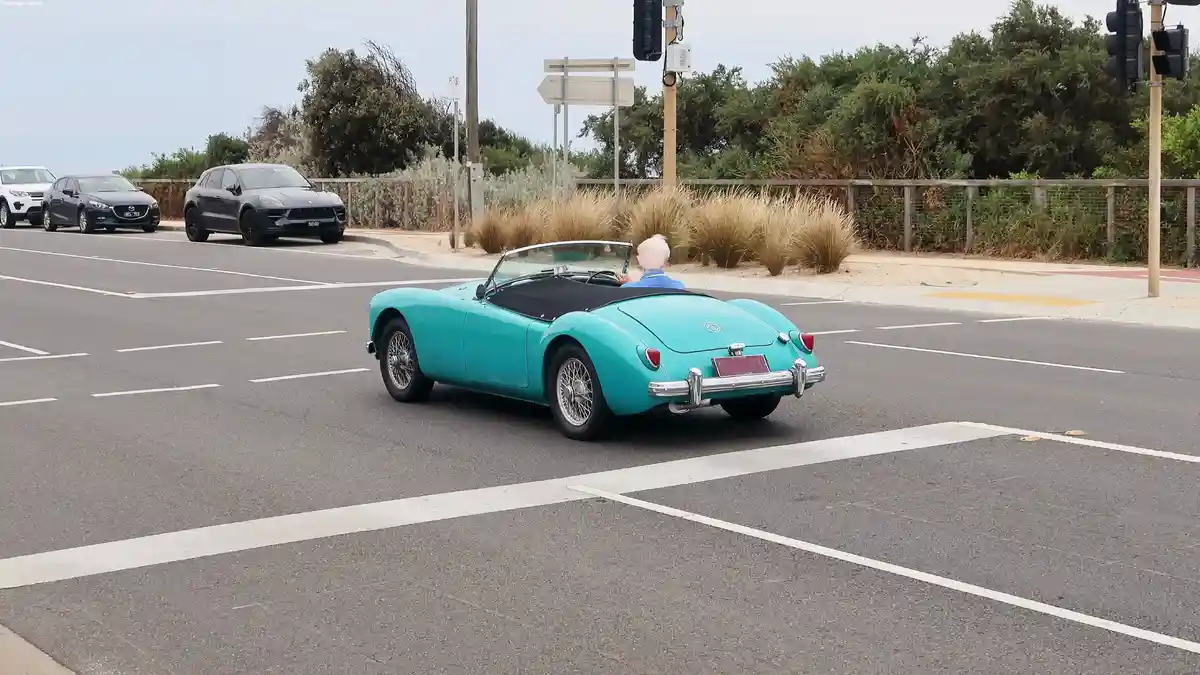 An elderly person with white hair drives a turquoise classic convertible MG sports car through a modern intersection, with…