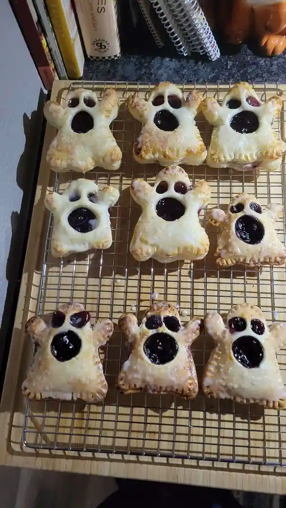 Nine baked pastries shaped like ghosts with dark jam filling for eyes and mouths sit on a cooling rack. The ghost-shaped…
