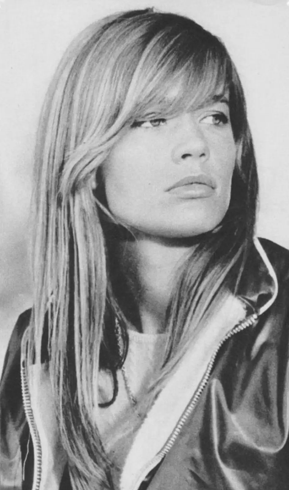 A black-and-white photo of a woman with long, straight hair and bangs, wearing a zip-up jacket, looking off to the side with…