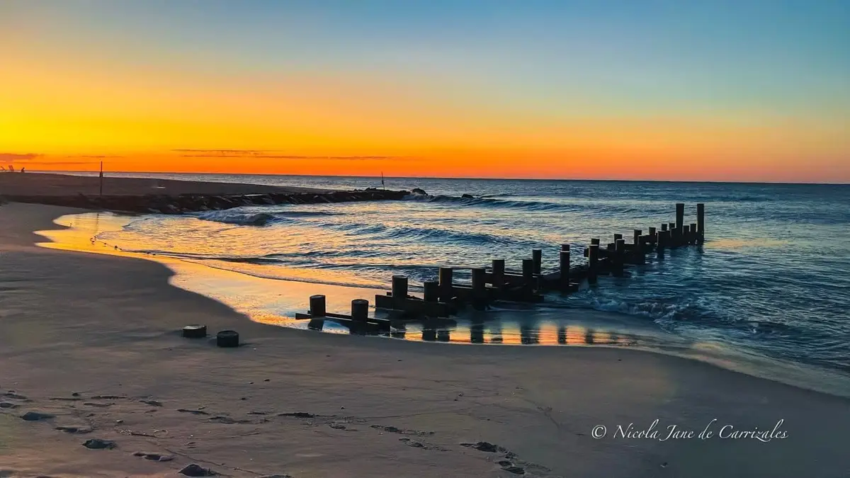 A serene beach sunset photo with orange and blue skies, gentle waves, and wooden pilings extending into the water. Two black…