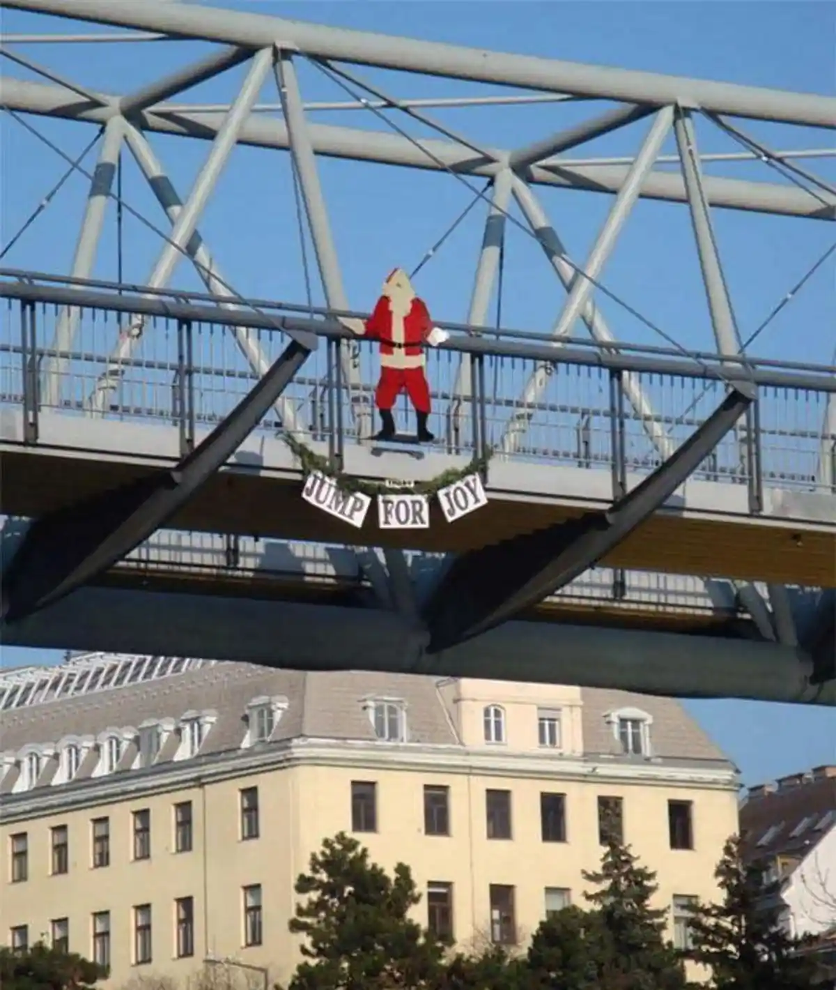 A large, life-sized Santa Claus cutout stands on a pedestrian bridge, holding onto the railing. Below him, a festive green…