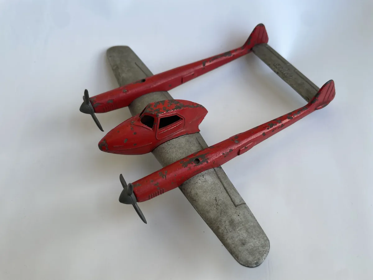 A vintage red and silver toy airplane with chipped paint and two propellers is lying on its back on a white surface. The…