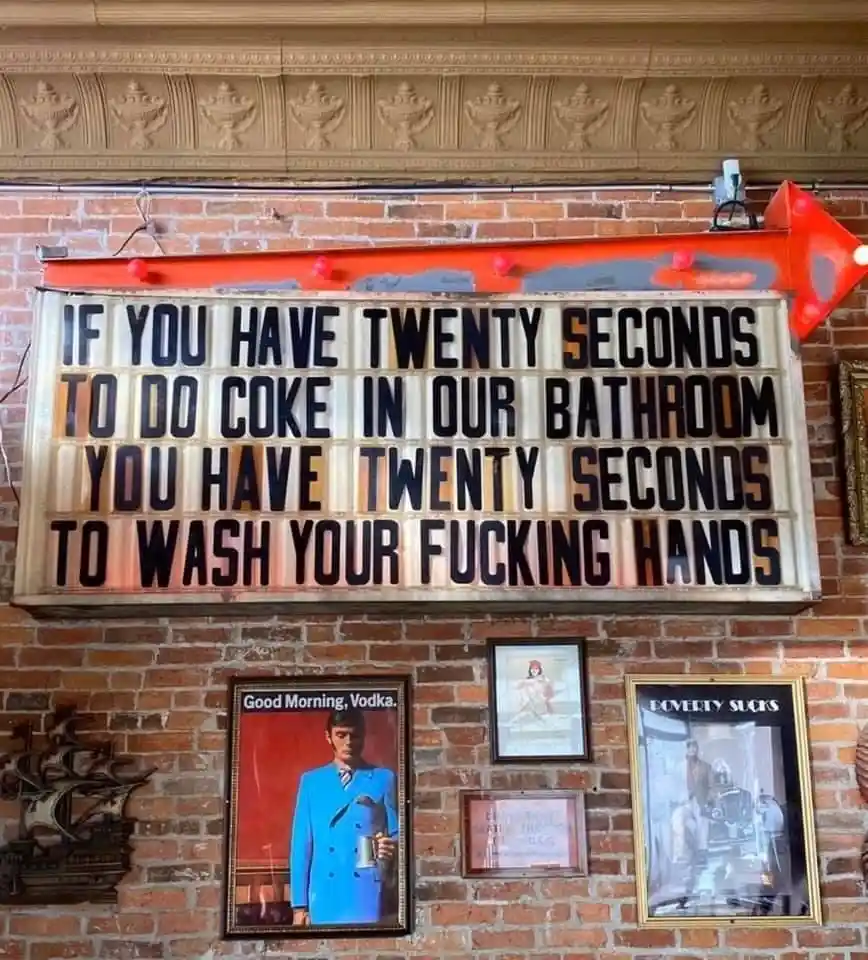 A weathered, illuminated marquee sign mounted on a brick wall reads: 'IF YOU HAVE TWENTY SECONDS TO DO COKE IN OUR BATHROOM…