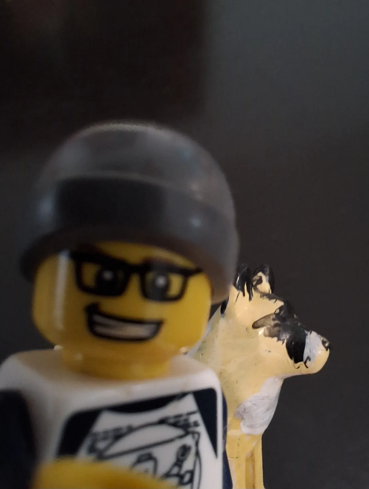 A close-up, slightly blurry photo shows a yellow Lego minifigure wearing a black helmet and glasses, smiling, with a crudely…