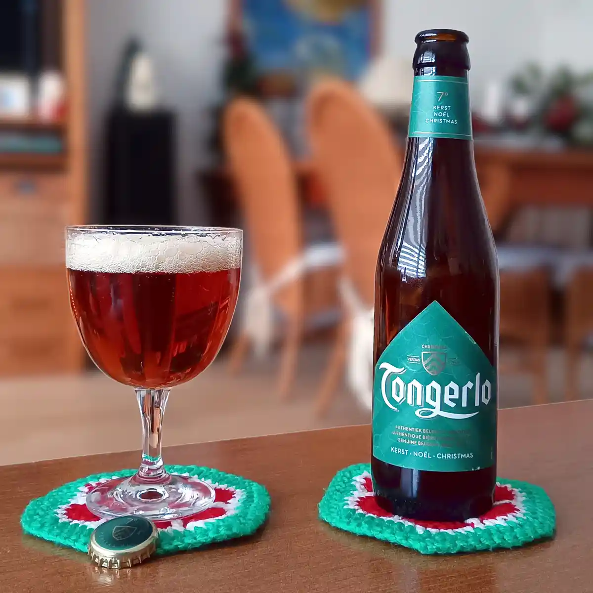 A bottle of Tangerlo Christmas beer and a filled glass sit on festive green, red, and white crocheted coasters on a wooden…