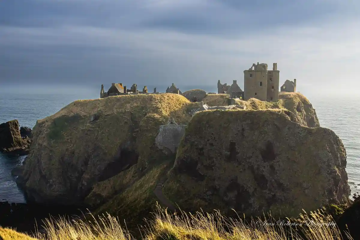 A scenic photograph of the ruins of a stone castle perched atop a grassy, rocky cliff overlooking the ocean under a cloudy…