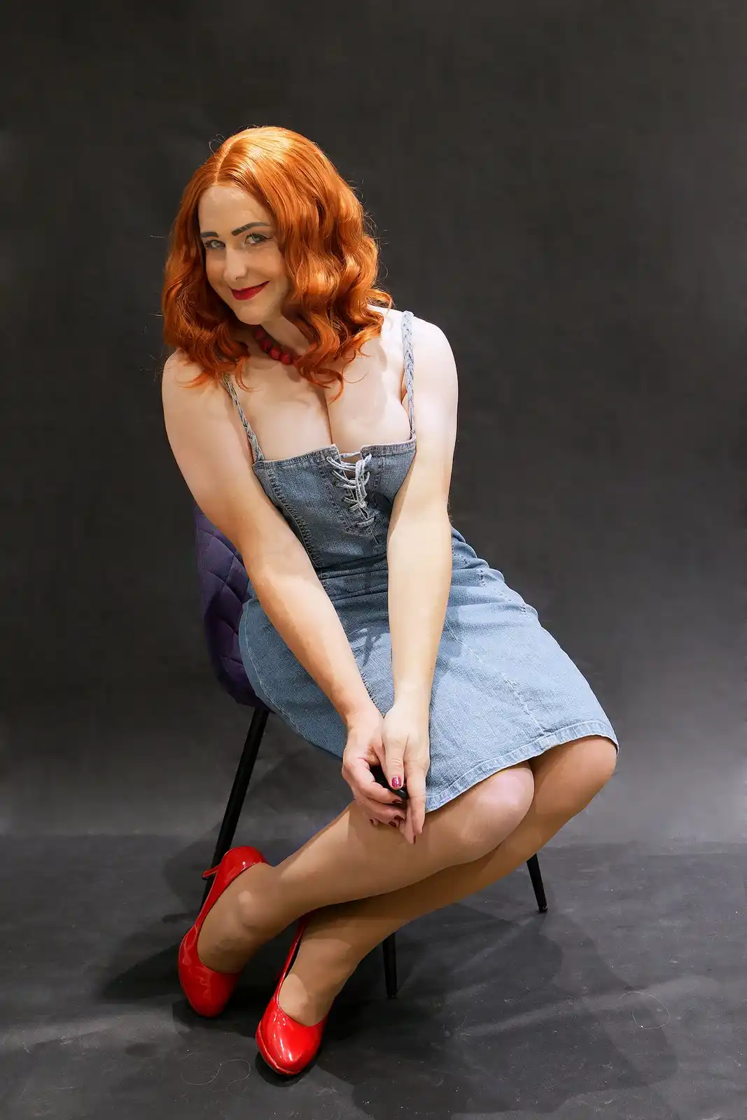 A woman with red hair and red lipstick sits on a purple chair wearing a denim dress with lacing at the bust and red patent…