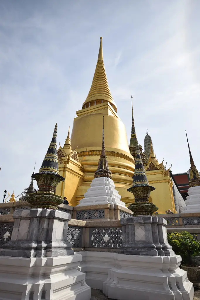 A low-angle photo of a grand golden temple with ornate spires under a partly cloudy sky, featuring white and grey stone…
