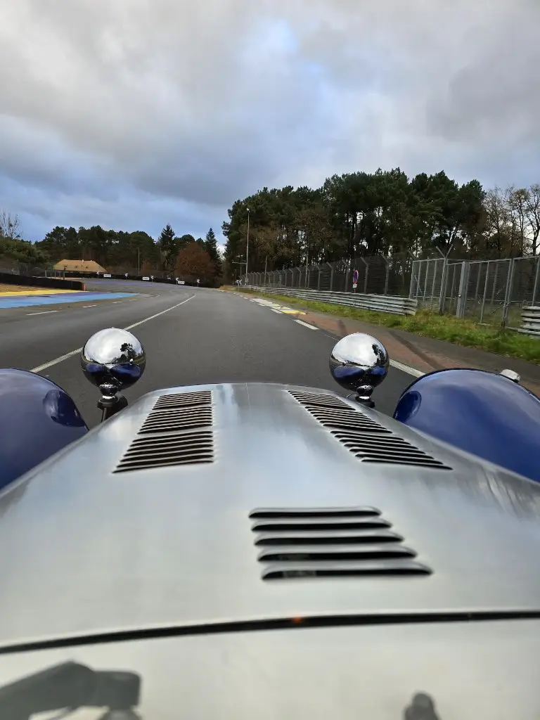 A first-person view from the hood of a vintage blue race car with chrome round headlights, driving on a racetrack under an…