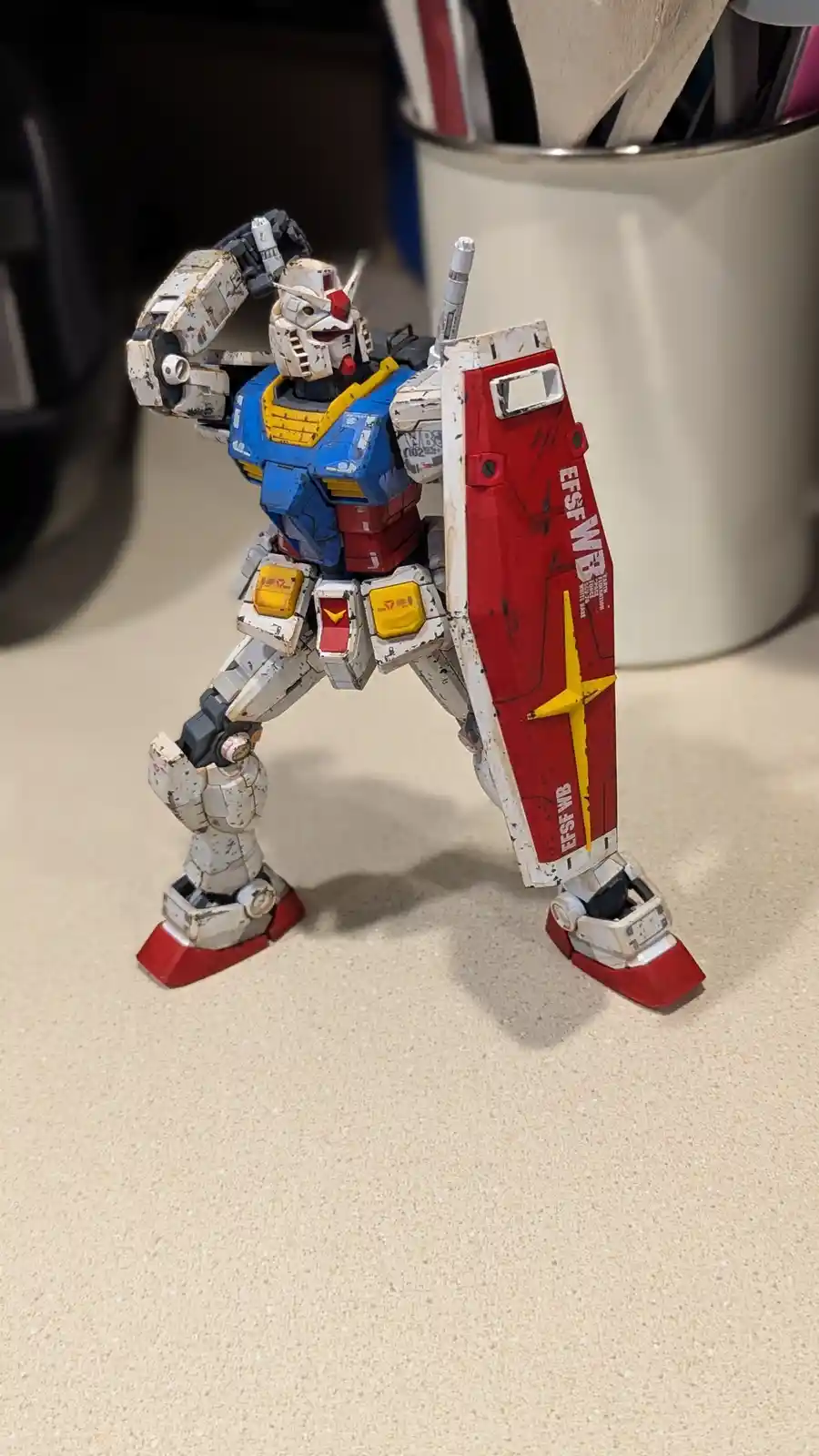 A weathered, battle-damaged blue, white, and red Gundam model stands on a beige countertop, holding a red shield with 'EFSF…