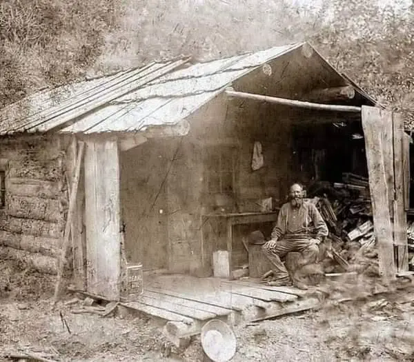 An old sepia-toned photograph shows a man sitting on a wooden porch in front of a rustic log cabin with a sloped roof. The…