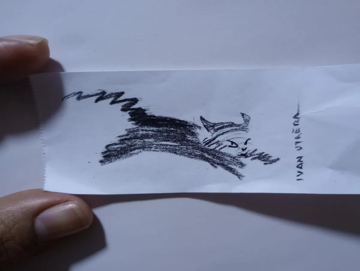A hand holds a small piece of paper with a crude, black crayon drawing of a cat-like creature with sharp teeth and a…