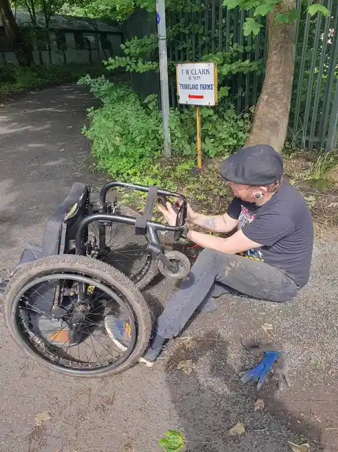 A man in a flat cap and jeans is kneeling on a gravel path, repairing a rugged, off-road wheelchair with thick tires. Beside…