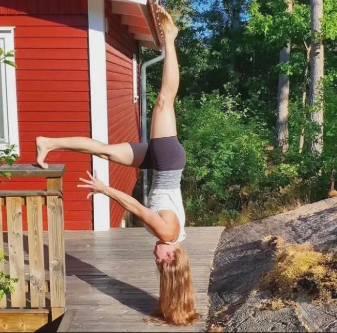 A woman with long blonde hair is performing a handstand on a wooden deck, balancing on her hands with her legs split in a…