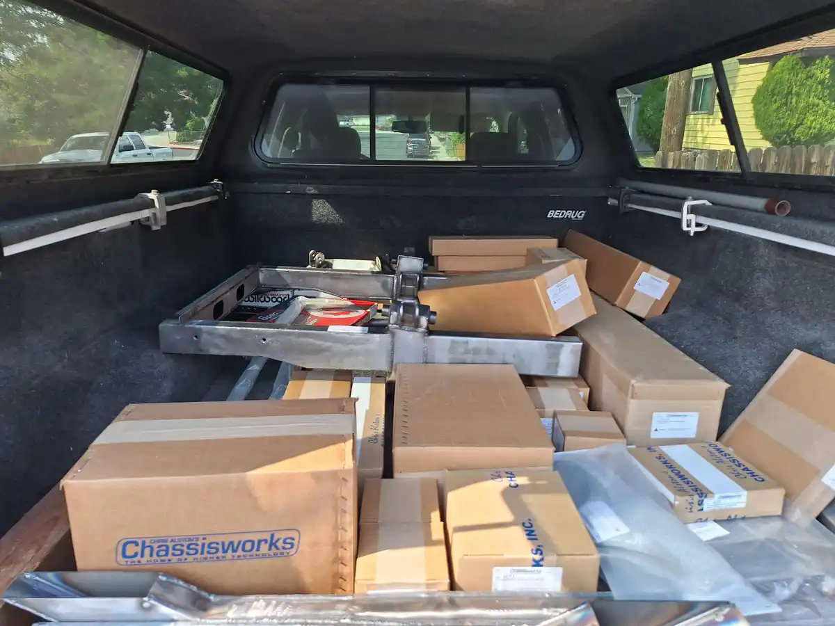 The bed of a pickup truck is filled with numerous cardboard boxes labeled 'Chris Alston's Chassisworks' and a large metal…