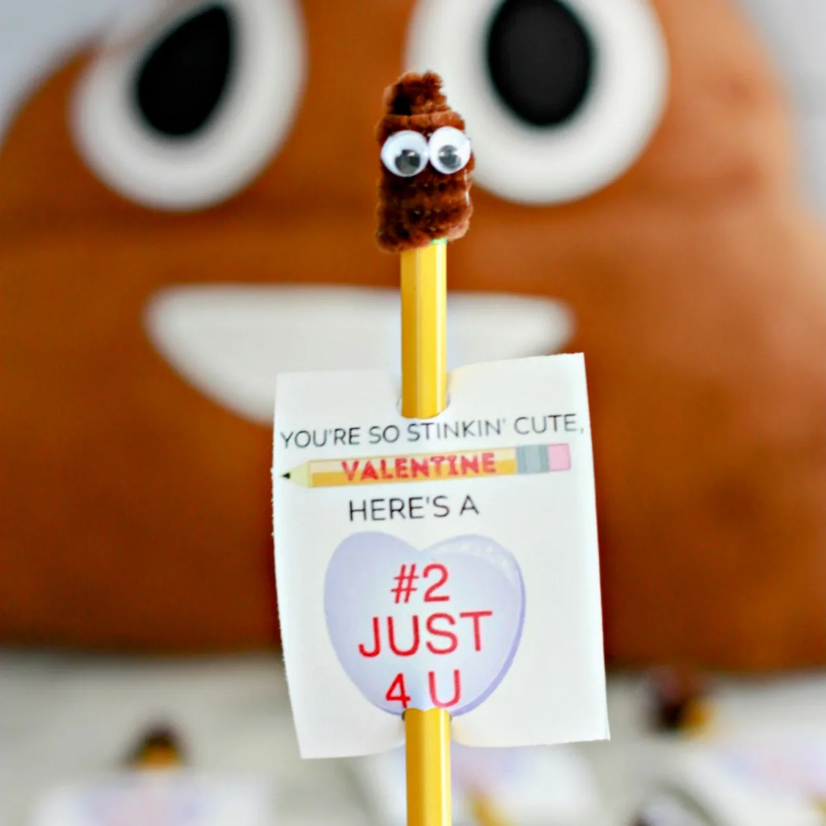 A handmade Valentine's Day card is attached to a yellow pencil, featuring a small brown fuzzy poop emoji with googly eyes on…