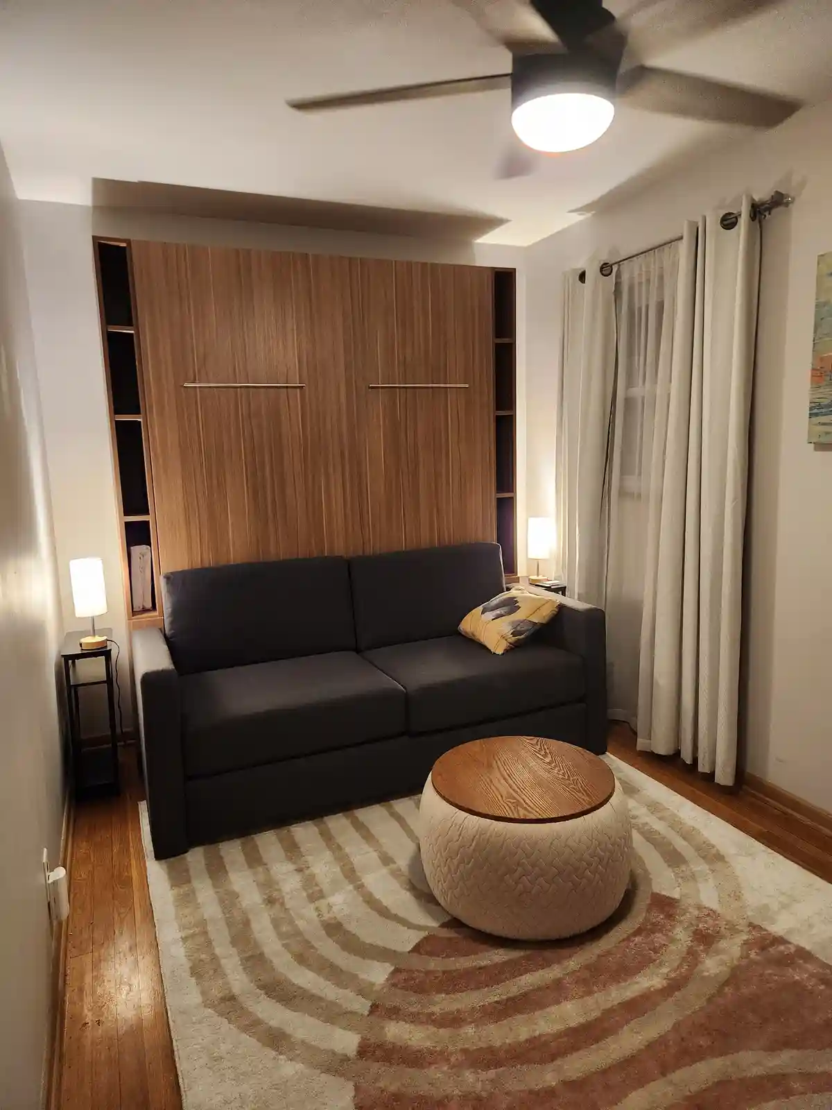 A cozy, modern living room features a dark gray sofa centered on a patterned rug, flanked by two lit table lamps on side…