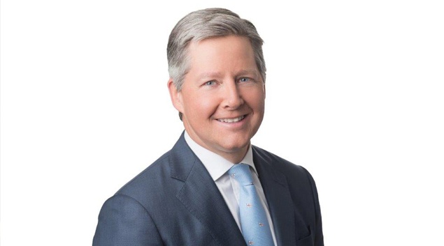 a professionally shot headshot of a smiling man with gray hair wearing a dark suit and light blue tie against a white…